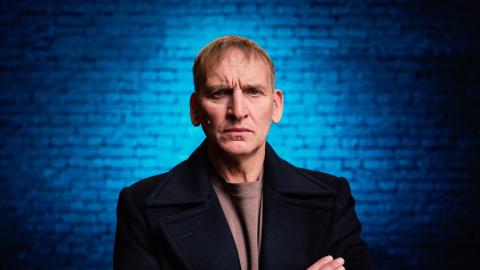 Christopher Eccleston standing with his arms crossed in front of a blue brick background